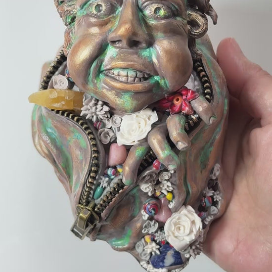 Hand made sculpture using Polymer clay, depicting a happy face exiting from a giant suit. This sculpture is painted to look like patinaed bronze and is decorated with natural stone, gems and sculpted flowers. 